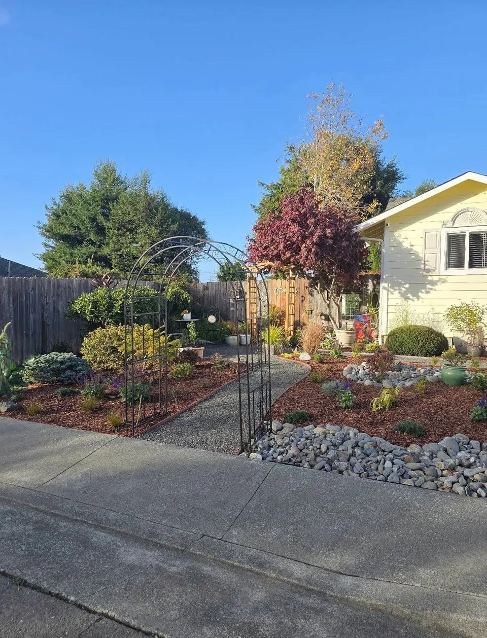 Front-yard cottage garden with an iron arbor, gravel path, river rock, and layered planting beds
