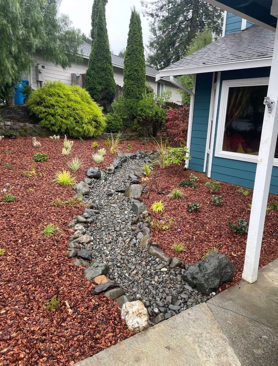 River-rock dry-creek drainage channel winding through red mulch beds and new plantings beside a home