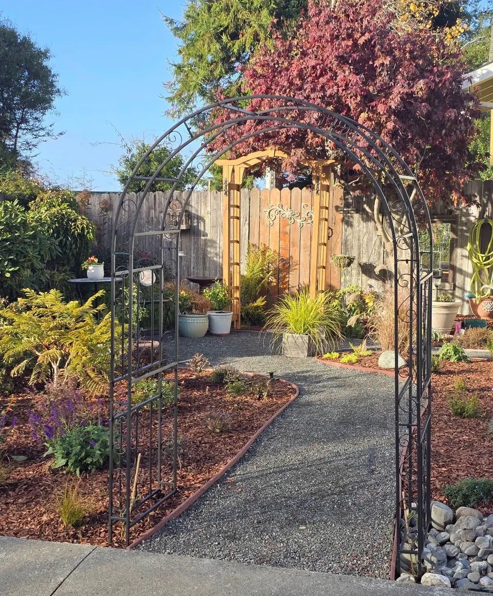 Iron garden arbor over a gravel path leading to a cedar gate, framed by fall color and potted plants