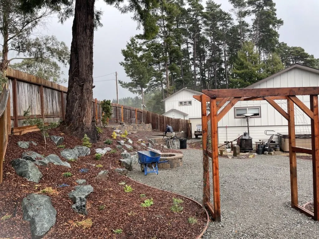 The same backyard slope finished with boulder planting beds, a block retaining wall, a paver fire pit, and a redwood arbor
