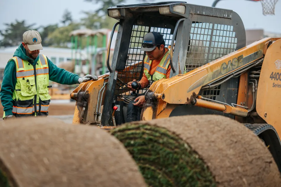 Landscaping crew in high-visibility vests loading a sod roll onto a skid steer