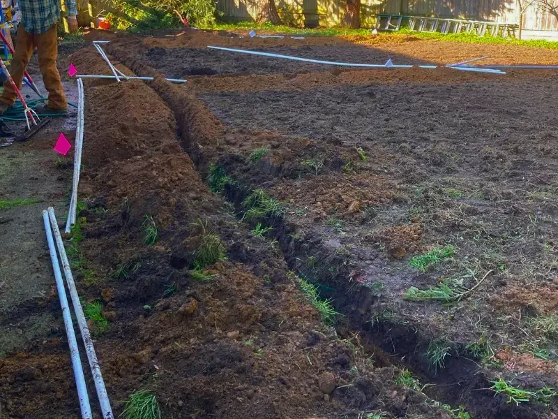 Trenched irrigation lines with pipe and marking flags across a bare yard